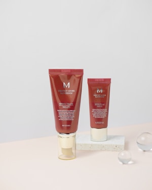 Two red cosmetic tubes labeled as BB cream are positioned vertically on a light-colored surface. Both tubes are marked with the brand name 'MISSHA' and they contain detailed product information. One tube is directly on the surface, while the other is placed on a white rectangular block. Clear glass spheres are scattered in the background, adding an aesthetic touch.
