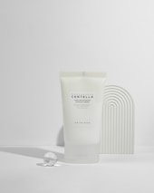A tube of Madagascar Centella tone brightening capsule cream is displayed against a minimalist background with soft shadows. Next to the tube is a small glass sphere and a textured curved object, creating a modern, clean aesthetic.