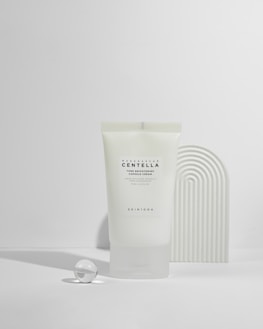 A tube of Madagascar Centella tone brightening capsule cream is displayed against a minimalist background with soft shadows. Next to the tube is a small glass sphere and a textured curved object, creating a modern, clean aesthetic.