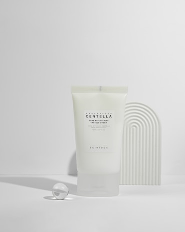 A tube of Madagascar Centella tone brightening capsule cream is displayed against a minimalist background with soft shadows. Next to the tube is a small glass sphere and a textured curved object, creating a modern, clean aesthetic.