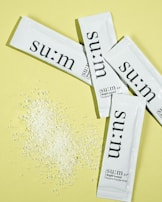 Several white sachets with the brand name 'su:m' printed on them are arranged on a pale yellow background. Near the sachets, there is a small pile of white granular powder.