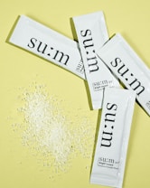 Several white sachets with the brand name 'su:m' printed on them are arranged on a pale yellow background. Near the sachets, there is a small pile of white granular powder.