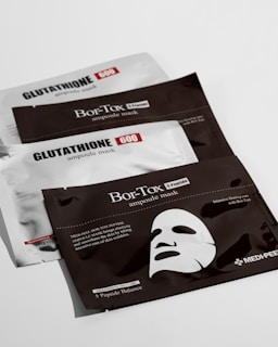 Three flat, rectangular packages are arranged on a white surface. Two of the packages are labeled 'GLUTATHIONE 600 ampoule mask' and are silver with black and red text. The middle package is labeled 'Bor-Tox ampoule mask' and is black with white text. The Bor-Tox package features a graphic of a facial mask and mentions skin elasticity and wrinkle care.