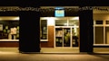 A storefront at night with holiday lights hanging above the entrance. The building is made of brick with large windows displaying various signs and posters inside. The signage above the door is illuminated.
