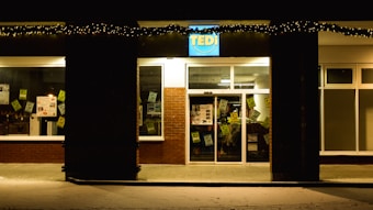 A storefront at night with holiday lights hanging above the entrance. The building is made of brick with large windows displaying various signs and posters inside. The signage above the door is illuminated.
