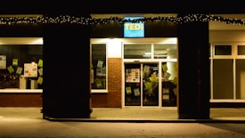 A storefront at night with holiday lights hanging above the entrance. The building is made of brick with large windows displaying various signs and posters inside. The signage above the door is illuminated.