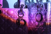 Two quilted designer handbags are illuminated under purple and magenta lighting. The material appears textured, with metallic and glossy surfaces reflecting light. Each bag features metallic rings and a decorative charm or logo.