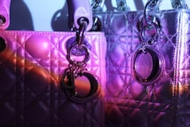 Two quilted designer handbags are illuminated under purple and magenta lighting. The material appears textured, with metallic and glossy surfaces reflecting light. Each bag features metallic rings and a decorative charm or logo.
