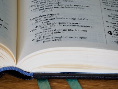 An open book laying on a wooden surface with visible text focusing on religious or biblical scripture. The book has a ribbon bookmark, and the pages display a mix of poetry-style verses and narrative text.