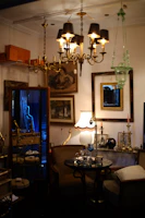 An antique store or vintage parlor setting with an assortment of ornate furniture and fixtures. Chandeliers with black lampshades hang from the ceiling, illuminating the room with a warm glow. The walls are adorned with framed art and mirrors. A small round glass table in the center has decorative items on it, surrounded by plush chairs. A lit table lamp with a beige, scalloped shade adds a cozy touch, while various brass candlesticks and decorative objects are placed on shelves and tables.