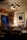 An antique store or vintage parlor setting with an assortment of ornate furniture and fixtures. Chandeliers with black lampshades hang from the ceiling, illuminating the room with a warm glow. The walls are adorned with framed art and mirrors. A small round glass table in the center has decorative items on it, surrounded by plush chairs. A lit table lamp with a beige, scalloped shade adds a cozy touch, while various brass candlesticks and decorative objects are placed on shelves and tables.