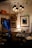An antique store or vintage parlor setting with an assortment of ornate furniture and fixtures. Chandeliers with black lampshades hang from the ceiling, illuminating the room with a warm glow. The walls are adorned with framed art and mirrors. A small round glass table in the center has decorative items on it, surrounded by plush chairs. A lit table lamp with a beige, scalloped shade adds a cozy touch, while various brass candlesticks and decorative objects are placed on shelves and tables.