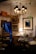 An antique store or vintage parlor setting with an assortment of ornate furniture and fixtures. Chandeliers with black lampshades hang from the ceiling, illuminating the room with a warm glow. The walls are adorned with framed art and mirrors. A small round glass table in the center has decorative items on it, surrounded by plush chairs. A lit table lamp with a beige, scalloped shade adds a cozy touch, while various brass candlesticks and decorative objects are placed on shelves and tables.