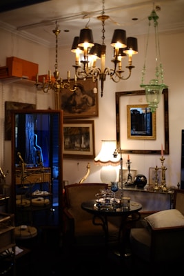 An antique store or vintage parlor setting with an assortment of ornate furniture and fixtures. Chandeliers with black lampshades hang from the ceiling, illuminating the room with a warm glow. The walls are adorned with framed art and mirrors. A small round glass table in the center has decorative items on it, surrounded by plush chairs. A lit table lamp with a beige, scalloped shade adds a cozy touch, while various brass candlesticks and decorative objects are placed on shelves and tables.