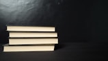Four neatly stacked hardcover books with a black cover are placed horizontally against a dark background. The edges of the pages create a contrasting light color with the black covers.