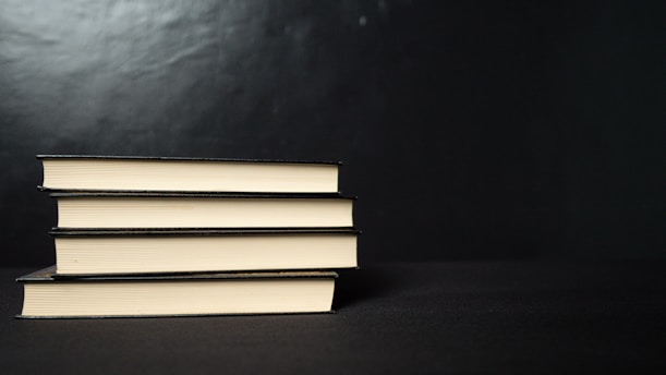 Four neatly stacked hardcover books with a black cover are placed horizontally against a dark background. The edges of the pages create a contrasting light color with the black covers.