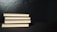 Four neatly stacked hardcover books with a black cover are placed horizontally against a dark background. The edges of the pages create a contrasting light color with the black covers.