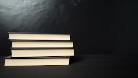 Four neatly stacked hardcover books with a black cover are placed horizontally against a dark background. The edges of the pages create a contrasting light color with the black covers.