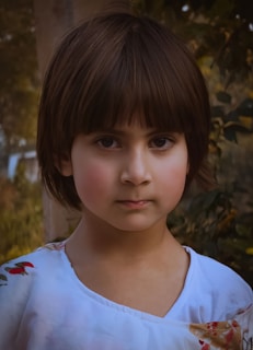 A young child with dark brown hair and rosy cheeks stands outdoors, wearing a white garment with a floral pattern on the shoulder. The background includes blurred greenery, suggesting a natural setting.