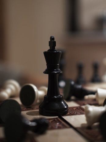 A black king chess piece stands prominently on a wooden chessboard surrounded by fallen white and black pieces. The surface of the board displays intricate patterns, while the background is softly blurred, highlighting the central figure.