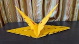 A handcrafted yellow paper sculpture resembling a geometric or abstract form is placed on a dark surface. The paper is intricately cut and folded, showing symmetry and precision. The background consists of curtains with vertical stripes in warm beige and brown tones, adding texture and depth.