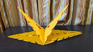 A handcrafted yellow paper sculpture resembling a geometric or abstract form is placed on a dark surface. The paper is intricately cut and folded, showing symmetry and precision. The background consists of curtains with vertical stripes in warm beige and brown tones, adding texture and depth.