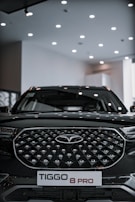 A sleek black SUV with a distinctive geometric grille pattern sits under a well-lit showroom ceiling. The vehicle prominently displays a badge in the center and a license plate reading 'TIGGO 8 PRO'.