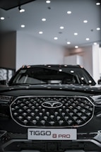 A sleek black SUV with a distinctive geometric grille pattern sits under a well-lit showroom ceiling. The vehicle prominently displays a badge in the center and a license plate reading 'TIGGO 8 PRO'.