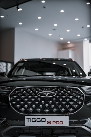 A sleek black SUV with a distinctive geometric grille pattern sits under a well-lit showroom ceiling. The vehicle prominently displays a badge in the center and a license plate reading 'TIGGO 8 PRO'.