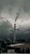 A tall industrial smokestack emits smoke against a backdrop of dark, cloudy skies. The surrounding area includes industrial buildings partially covered with snow. Distant hilltops are visible through the mist, enhancing the gloomy atmosphere.