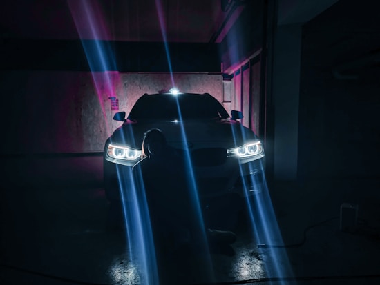 A car with illuminated headlights is parked in a dimly lit garage. Beams of light create a dramatic effect across the dark surroundings. A person is sitting or crouching in front of the car, mostly in silhouette.