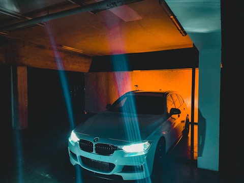 A white car with illuminated headlights is parked in a dimly lit underground garage. The surrounding environment is dark, with a stark contrast created by vivid orange and blue light streaks. The car is positioned near a concrete pillar with visible pipes on the ceiling, adding to the urban, industrial feel of the scene.