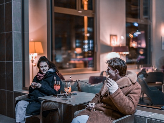 Two people are sitting in a cozy, dimly lit indoor setting. They appear to be enjoying each other's company, with drinks placed on a small table between them. The ambiance is warm, enhanced by soft indoor lighting, and there are reflections of lights in the background.