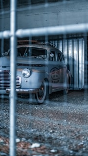 A vintage car is parked inside a gritty, industrial-looking garage or storage unit. The vehicle has weathered paint and a surf shop logo on its side. The image is taken through a wire fence, adding a sense of separation or confinement.