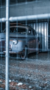 A vintage car is parked inside a gritty, industrial-looking garage or storage unit. The vehicle has weathered paint and a surf shop logo on its side. The image is taken through a wire fence, adding a sense of separation or confinement.