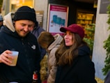 A group of people socializing outdoors. One person is holding a beverage in a plastic cup and wearing a black jacket and beanie. Another person, wearing a red cap and dark coat, is smiling and appears to be engaged in conversation. The background shows a storefront or market setting with a festive atmosphere.