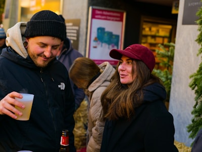 A group of people socializing outdoors. One person is holding a beverage in a plastic cup and wearing a black jacket and beanie. Another person, wearing a red cap and dark coat, is smiling and appears to be engaged in conversation. The background shows a storefront or market setting with a festive atmosphere.