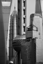 A black and white photograph showcasing a cluster of modern skyscrapers with sleek, geometric designs. The architecture features various shapes including cylindrical pillars and spherical elements, conveying an urban, futuristic aesthetic.