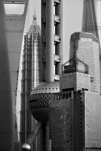 A black and white photograph showcasing a cluster of modern skyscrapers with sleek, geometric designs. The architecture features various shapes including cylindrical pillars and spherical elements, conveying an urban, futuristic aesthetic.