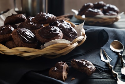 A wicker basket filled with chocolate-covered cookies is placed on a dark cloth on top of a wooden table. In the background, there is another basket with similar cookies. Several pieces of cutlery, including spoons and forks, are scattered on the table next to the basket. One cookie is broken into pieces and placed on the tablecloth.
