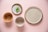 A collection of four ceramic dishes arranged on a light pink background. The dishes vary in size and finish, showing earthy tones with textures. The largest plate is plain with a speckled design, while the small dish holds a fresh green sprig.