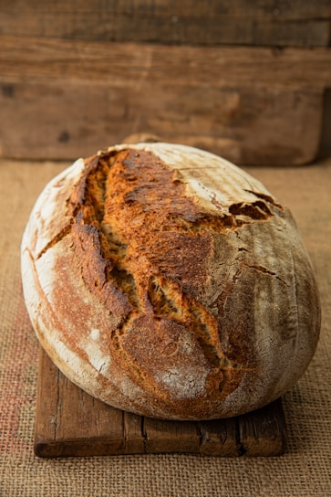 A rustic loaf of bread with a golden-brown crust sits on a wooden board against a burlap background. The bread is oval-shaped with visible scoring on the top, giving it an artisanal appearance.