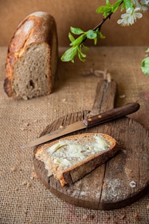 A rustic setting with a slice of brown bread topped with butter placed on a round wooden cutting board. A knife lays next to the slice, and a whole loaf of bread is positioned in the background. Fresh green leaves and white blossoms hang from above, adding a touch of nature.