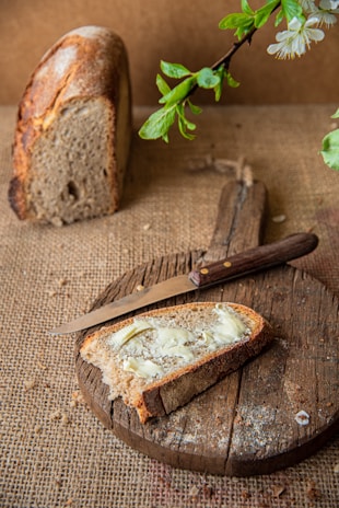 A rustic setting with a slice of brown bread topped with butter placed on a round wooden cutting board. A knife lays next to the slice, and a whole loaf of bread is positioned in the background. Fresh green leaves and white blossoms hang from above, adding a touch of nature.
