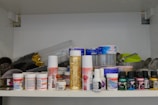 A shelf filled with various art supplies, including spray cans, jars of paint, and bottles of gel. The items are arranged in an organized manner, showcasing a range of brands and types. There are also some metal containers and small tools in the background.