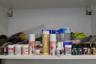 A shelf filled with various art supplies, including spray cans, jars of paint, and bottles of gel. The items are arranged in an organized manner, showcasing a range of brands and types. There are also some metal containers and small tools in the background.