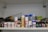 A shelf filled with various art supplies, including spray cans, jars of paint, and bottles of gel. The items are arranged in an organized manner, showcasing a range of brands and types. There are also some metal containers and small tools in the background.