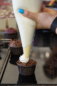 A hand is holding a piping bag with cream, decorating a chocolate muffin on a reflective surface. The hand has blue nail polish and a black wristband.