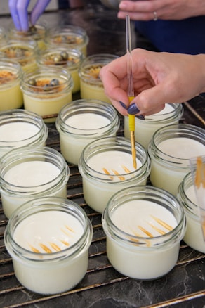 Hands are applying a decorative touch to creamy desserts in small glass jars using a pipette. The jars are arranged on a cooling rack, and some have additional toppings like nuts or berries. One hand is holding the pipette with yellow liquid, creating lines or patterns on top of the desserts.