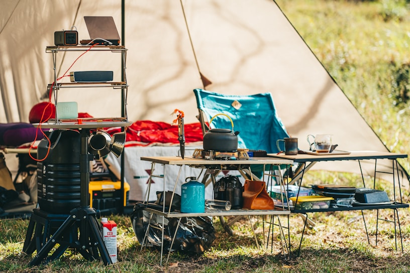 A campsite setup with various items including a kettle on a portable stove, a folding chair, and multiple tables holding kitchen equipment and supplies. There is a shelf unit with gadgets including a laptop and speakers, and an energy device with cables attached. A tent provides shade in the background, with grass underfoot.